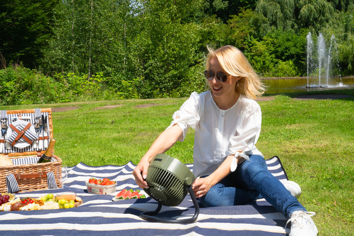 Eine Frau mit Sonnenbrille sitzt bei einem Picknick auf einer gestreiften Decke und greift nach dem tragbaren Unold Akkuventilator Outdoor. Ein Picknickkorb mit Essen steht daneben, und ein Brunnen mit Grünpflanzen umgibt die Szene.