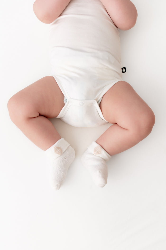 A baby lying on their back wearing a white bodysuit and white socks on a clean white background.