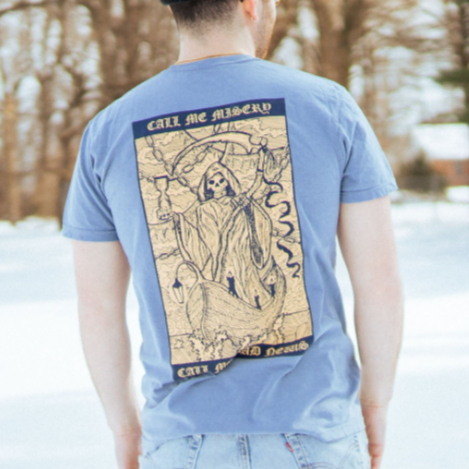 Back of Blue Jean colored short sleeve shirt with rectangle across the back. Inside the rectangle is a creature similar to the grim reaper riding in a wooden boat on the water. On top of that, there is text that says CALL ME MISERY. On the bottom of the image, there is text that says CALL ME BAD NEWS. There is a model wearing the shirt outside, with snow on the ground.