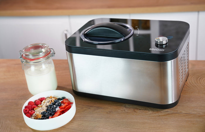Die Unold Eismaschine & Joghurtbereiter EVA steht auf einer hölzernen Arbeitsplatte neben einem Glas Milch und einer Schüssel mit Müsli, frischen Beeren und geschnittenen Erdbeeren.