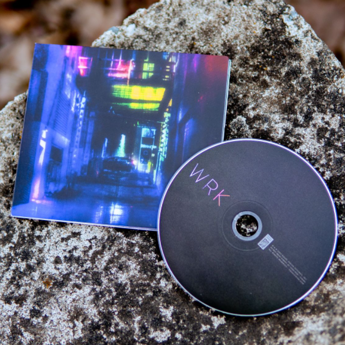 Square CD with artwork of an alley. The entire album cover is multicolored pink, dark blue, purple, and green. At the end of the alley there is a dark with its rear lights on. To the right of the album cover is a black CD with text that says WRK on the left hand side, colored in the same way as the cover. Both are laying on top of a granite surface.