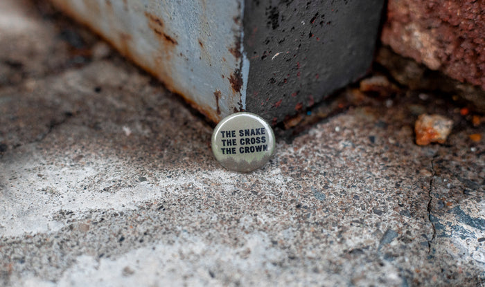 A brown pin button featuring the brand name "THE SNAKE THE CROSS THE CROWN" in bold black uppercase letters rests on rough concrete by a metal and brick corner.