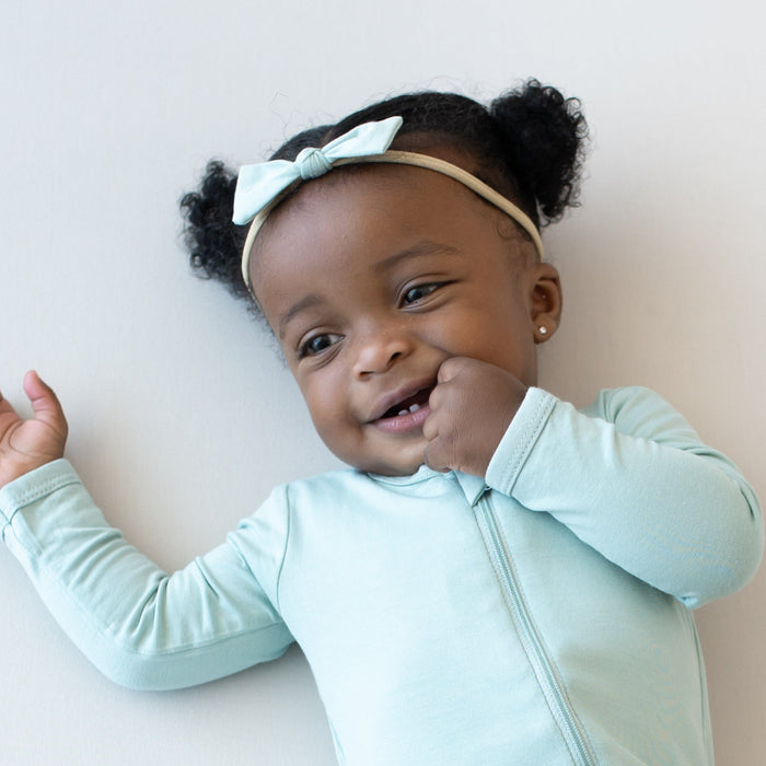 Infant wearing a Kyte Baby Tiny Bow and Zippered Romper in Sage with one hand in mouth
