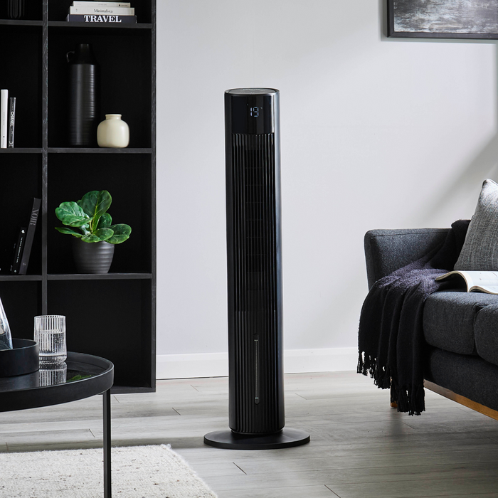 Image shows off the 3-in-1 black Geepas air cooler tower fan in the middle of a living room with furniture around it.