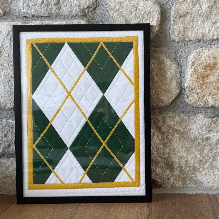 Framed quilt with green, white, and gold pattern against a stone wall.
