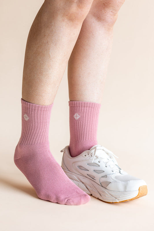 A close-up of a person's feet wearing pink crew socks and a white athletic sneaker against a solid light beige background.