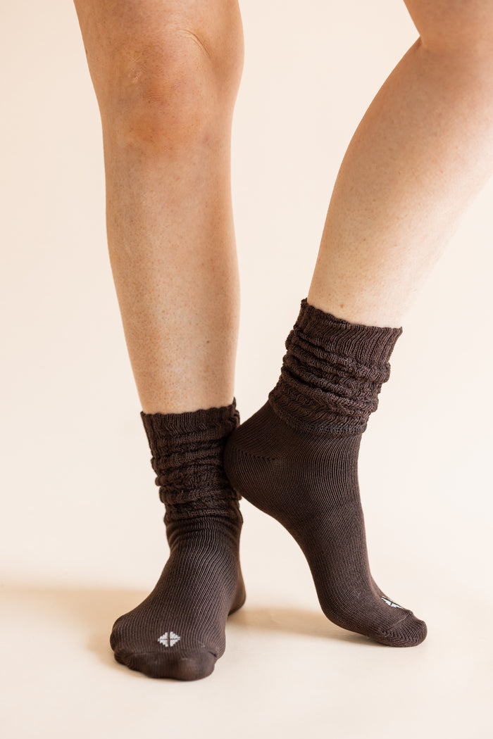 A close-up of a person's legs wearing dark brown ribbed crew socks against a solid light beige background.