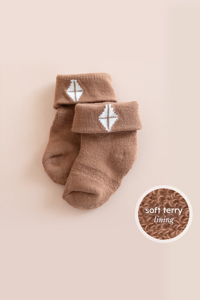 A pair of soft brown baby socks with a white geometric pattern on the cuff, displayed on a light beige background with a circular inset showing the soft terry lining texture.