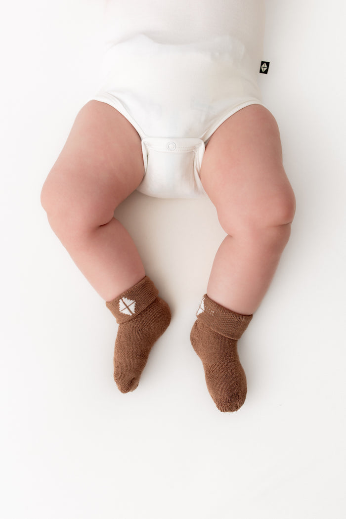 A baby's legs wearing brown knit socks with a small white geometric logo, shown from a top-down view on a white background.