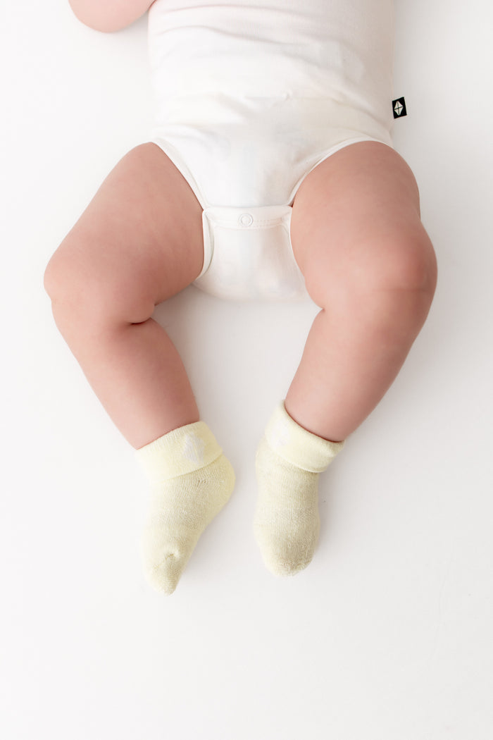 A baby's legs wearing pale yellow ankle socks and a white bodysuit, photographed from above on a clean white background.