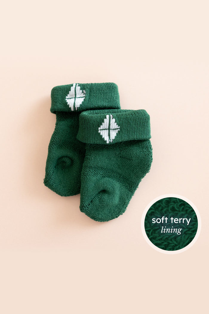 A pair of dark green baby socks with white knit geometric patterns on the cuffs, displayed on a light peach background with a circular inset showing the soft terry lining texture.
