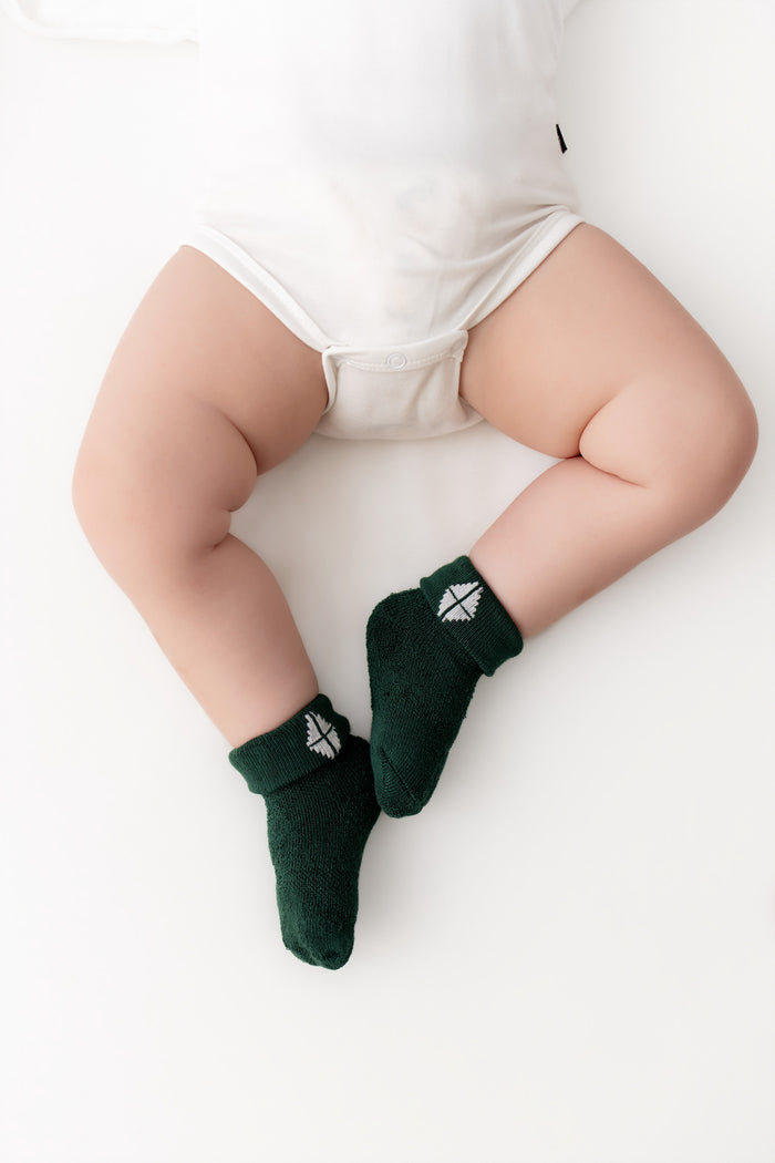 A baby's legs wearing dark green knit socks with a small white geometric logo, styled with a white bodysuit on a clean white background.