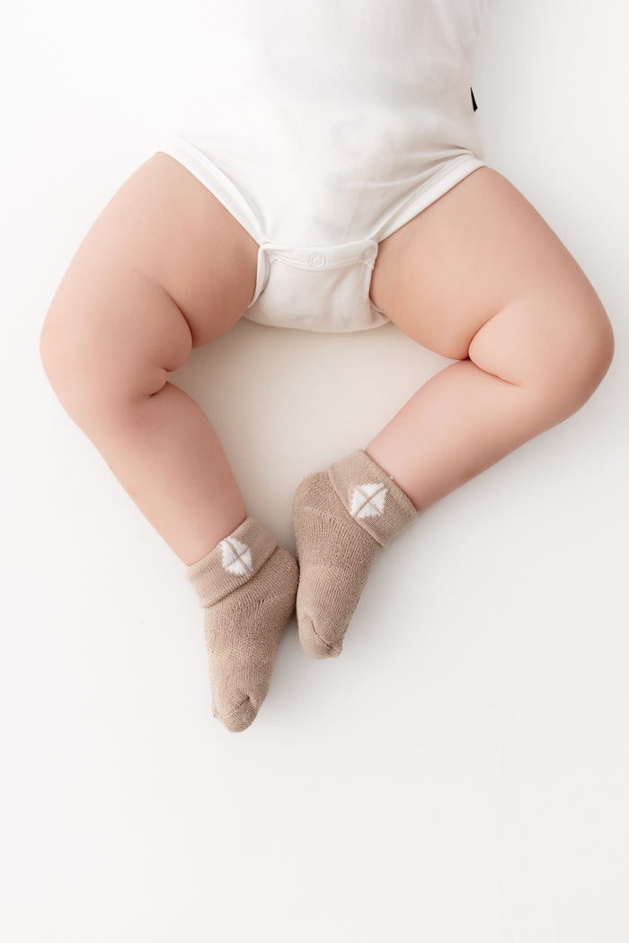 A baby's legs wearing beige ankle socks with a small white geometric logo, styled with a white bodysuit on a clean white background.