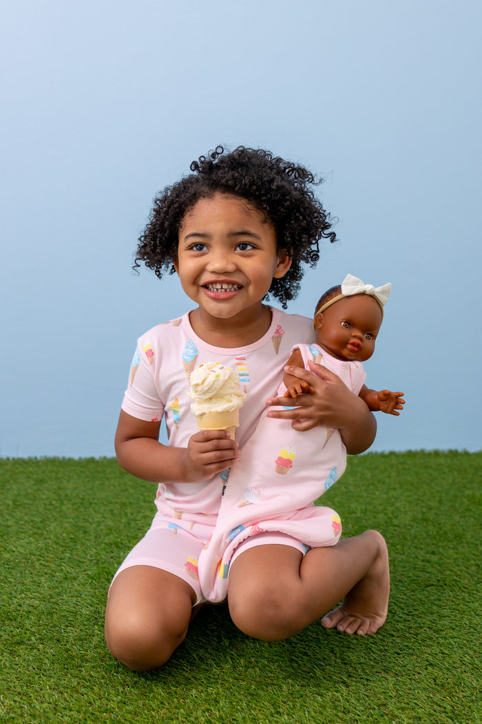 Child holding Baby doll sleep bag in Sakura Ice Cream print wearing a similar print Short sleeve pajama set sitting on green grass with an ice cream cone