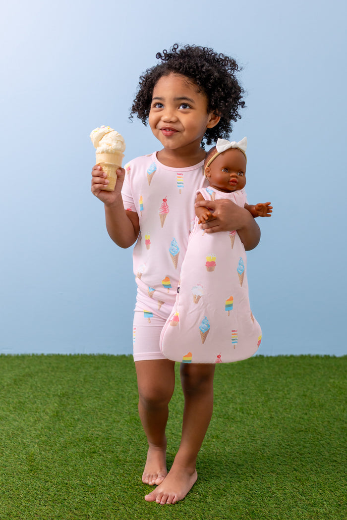 Child holding Baby doll sleep bag in Sakura Ice Cream print wearing a similar print Short sleeve pajama set with ice cream cone on green grass