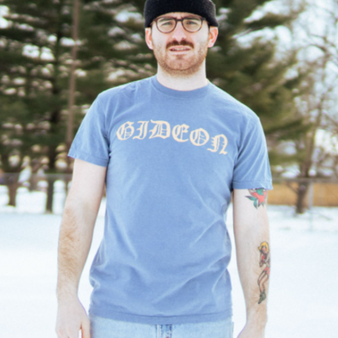 Blue Jean colored short sleeve shirt with yellow text across the chest that says GIDEON. There is a model wearing the shirt standing outside with snow on the ground.