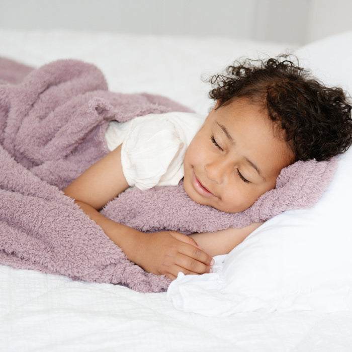 A little girl lays under a Bamboni® blanket by Saranoni. This ultra-soft blanket is a light purple blanket and made from a premium blend of bamboo rayon and poly microfiber, offering a lightweight, breathable, and fluffy feel. Perfect as a toddler blanket, luxury blanket, or soft blanket, it adds warmth and elegance to any bedroom and is a kids blanket. Designed for ultimate comfort, this buttery-soft Saranoni blanket is ideal for gifting. Enjoy this cozy blanket for years to come!