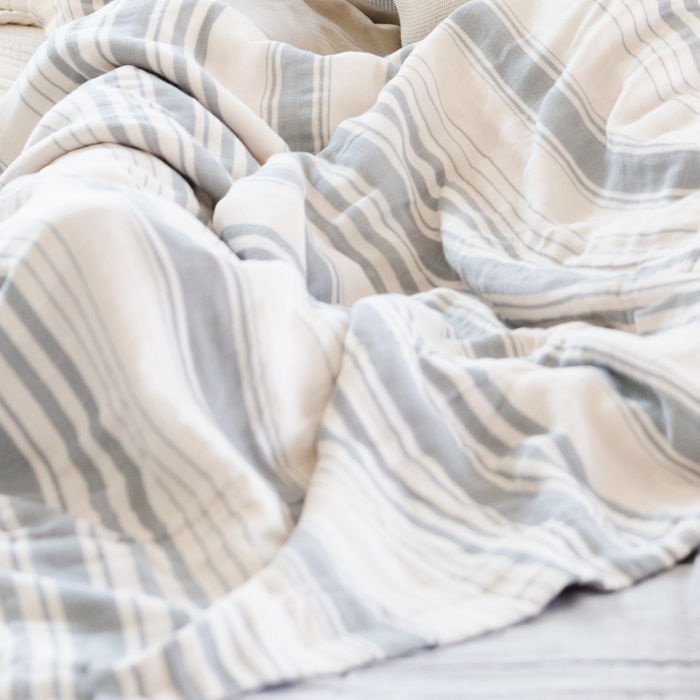 A blue and white Saranoni Queen/King muslin quilt in a soft stripe print. This luxury bedding blanket features four layers of ultra-soft bamboo muslin, creating an airy yet cozy feel. Designed with a stitched quilted pattern and a one-inch border, this breathable blanket is perfect for muslin lovers looking for extra warmth and comfort. This luxury blanket is the softest blanket and a Saranoni blanket.
