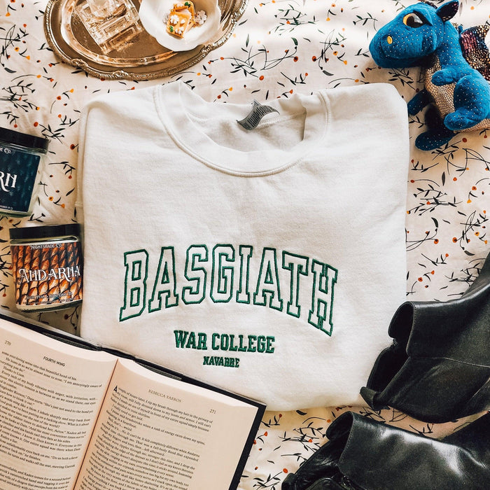 White Basgiath War College embroidered sweater folded on patterned fabric, surrounded by book, candles, and plush dragon.