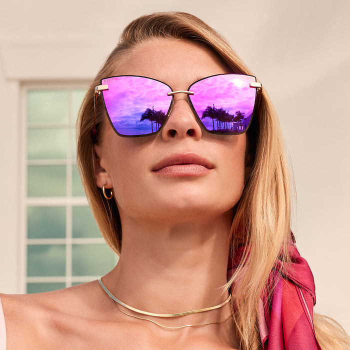 lifestyle - female outdoors wearing diff eyewear featuring the becky v cat eye sunglasses with a gold frame and pink rush mirror lenses front view with reflection of the sky in the lenses