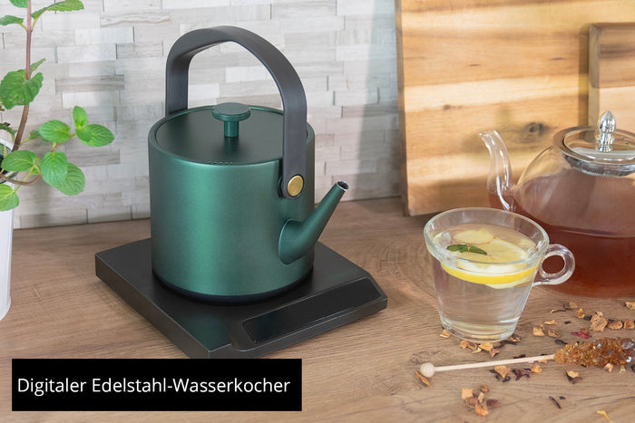 Der Unold Blitzkocher Jade, ein grüner digitaler Edelstahl-Wasserkocher, steht auf einem schwarzen Sockel neben einer Glasteekanne, einer Tasse Zitronentee, losen Teeblättern und einer Topfpflanze. Der Text lautet: „Digitaler Edelstahl-Wasserkocher“.