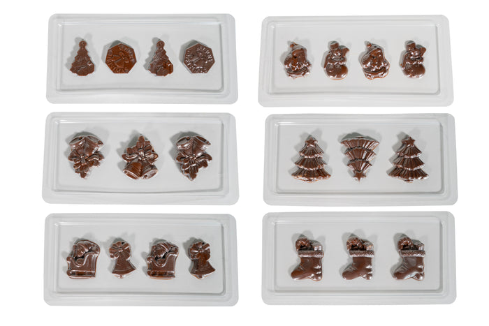 Sechs weiße Tabletts aus der Chocolatier Weihnachtsedition von Unold zeigen glänzende Schokoladenstücke in Form von Weihnachtsbäumen, Strümpfen, Glocken, Schneemännern und anderen festlichen Weihnachtsmotiven auf weißem Hintergrund.