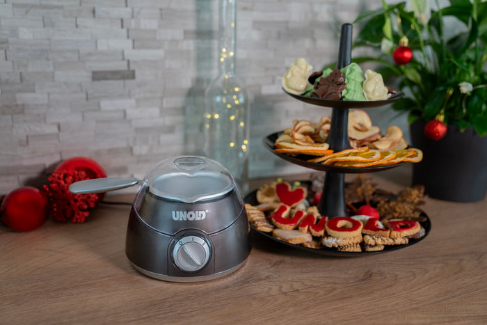 Die Schokoladenschmelzmaschine „Weihnachtsedition“ von Unold Chocolatier steht neben einem schwarzen, zweistöckigen Tablett, gefüllt mit verschiedenen Keksen, Trockenfrüchten und Pralinen. Weihnachtsschmuck und eine Pflanze schmücken den Hintergrund.