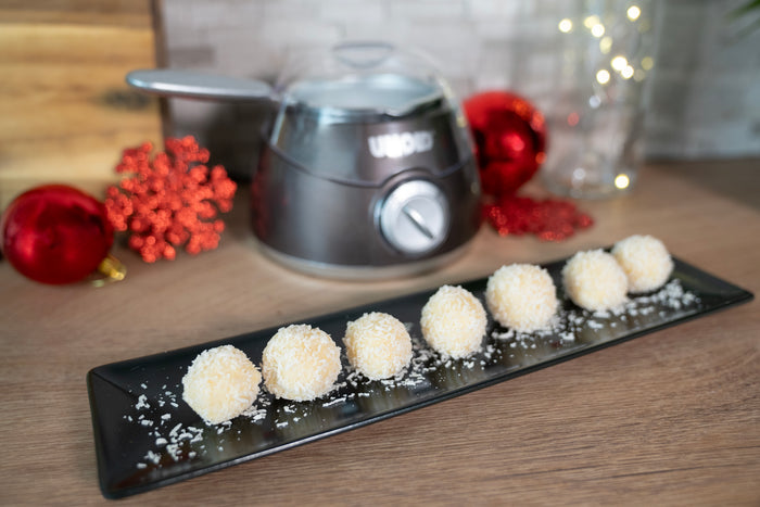 Ein schwarzer rechteckiger Teller mit sieben Kokosnusskugeln aus der Unold Chocolatier Weihnachtsedition steht auf einer Holzfläche. Im Hintergrund sind rote Ornamente, ein silbernes Unold-Gerät und dekorative Lichter zu sehen.