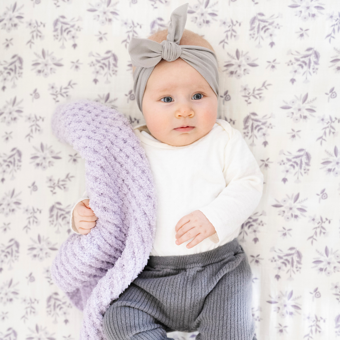 A baby girl holds a light purple waffle knit blanket. The soft blanket is a baby blanket and a Saranoni blanket. She is laying on top of a white crib sheet with a light purple floral design on the crib sheet. This crib sheet is a soft crib sheet and is a Saranoni crib sheet.