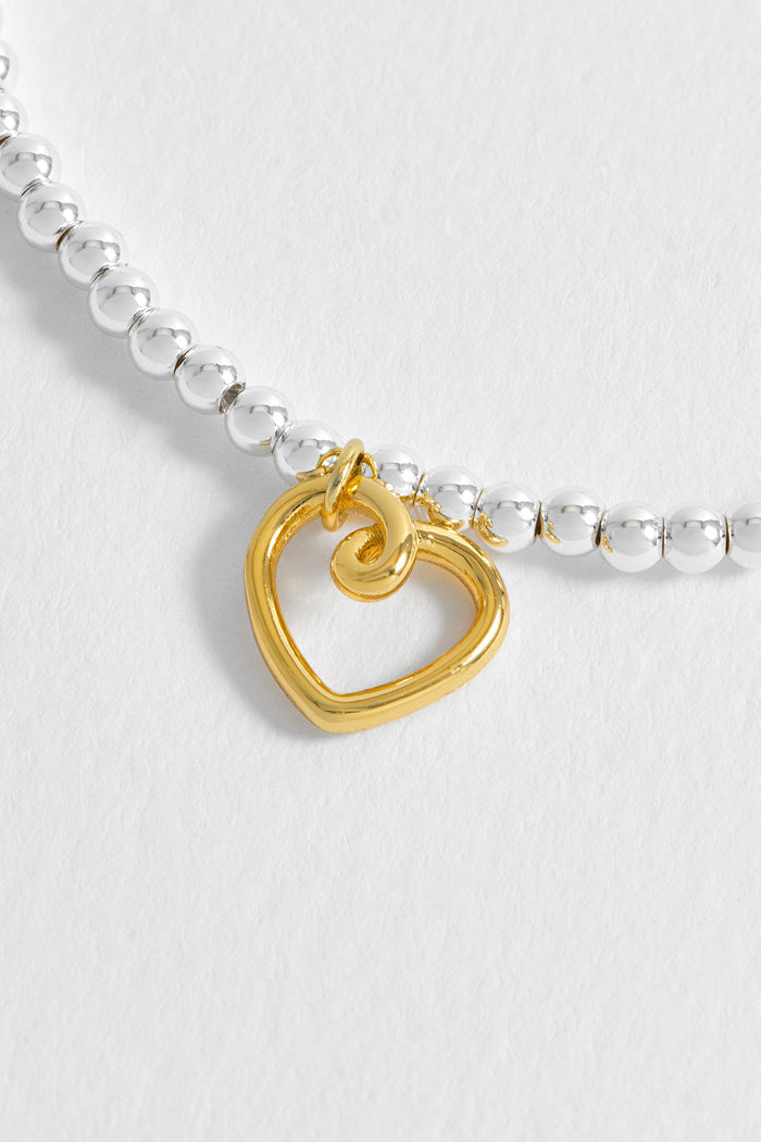 Gold heart-shaped charm on a silver beaded bracelet against a white background