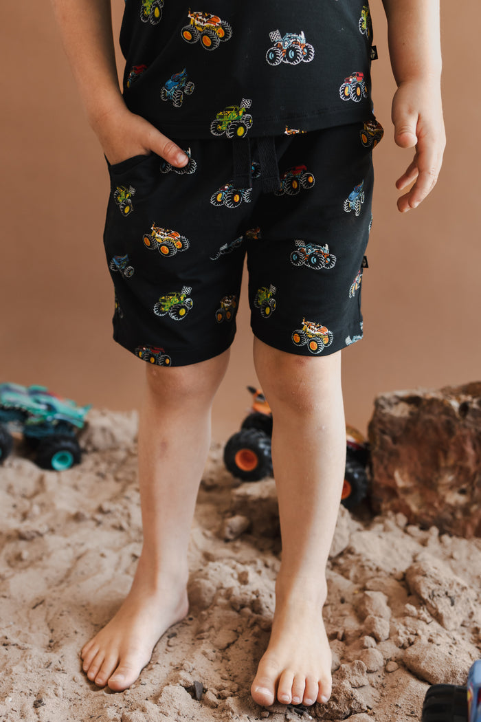 Boy wearing Black drawstring short in Monster Truck print on sand and rocks with toy trucks