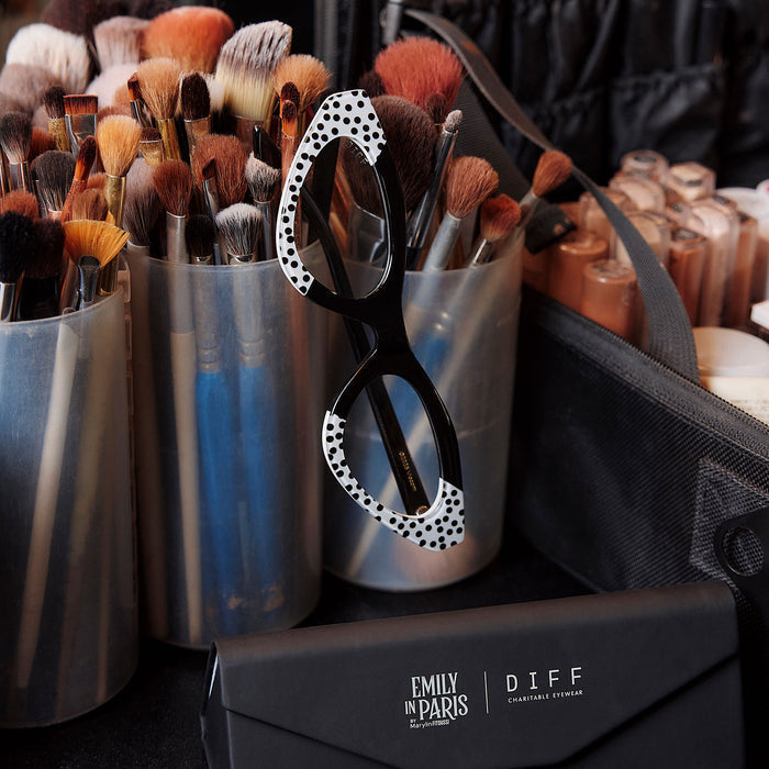 still life of emily in paris x diff eyewear featuring the emily 2.0 cat eye prescription glasses with a midnight polka dot frame surrounded by makeup brushes