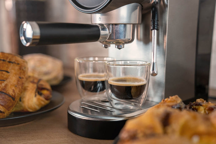 Eine Nahaufnahme der Unold Espressomaschine Piccopresso, die zwei Shots in Glastassen brüht, mit Tellern mit Gebäck, die im Vordergrund und Hintergrund auf einer Holzoberfläche angeordnet sind.