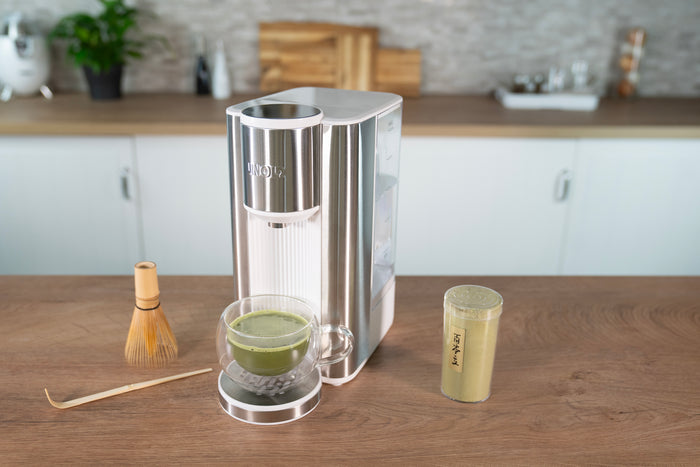 Der Unold Express Heißwasserbereiter Ava in modernem Weiß steht auf einer hölzernen Küchentheke. Daneben stehen ein Glas grüner Matcha, ein Bambusbesen, ein Löffel und ein Plastikbecher mit eisgekühltem Matcha. Im Hintergrund eine aufgeräumte Küche.