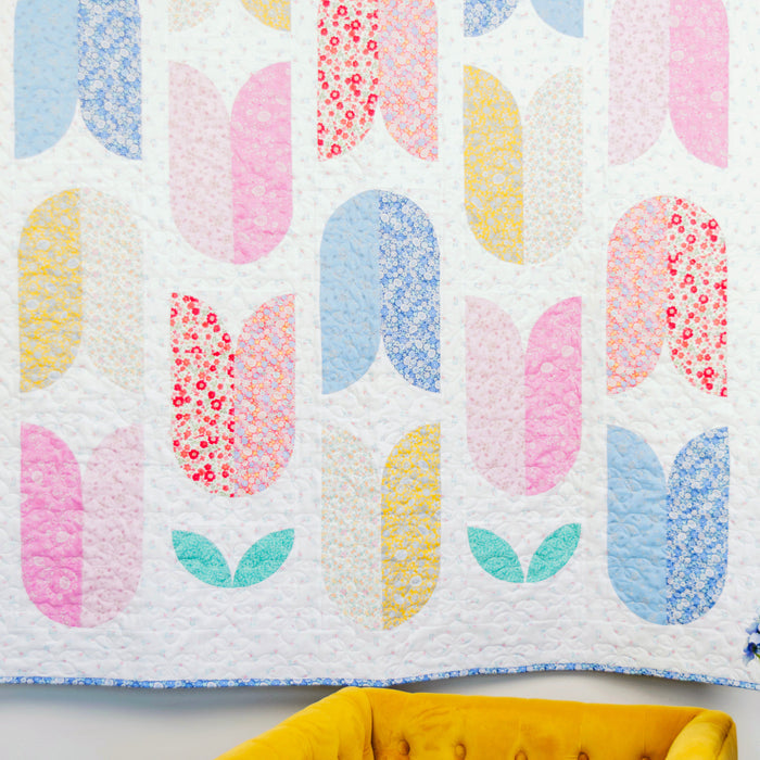 Colorful quilt with abstract pattern on a white wall with a yellow chair and blue flowers.