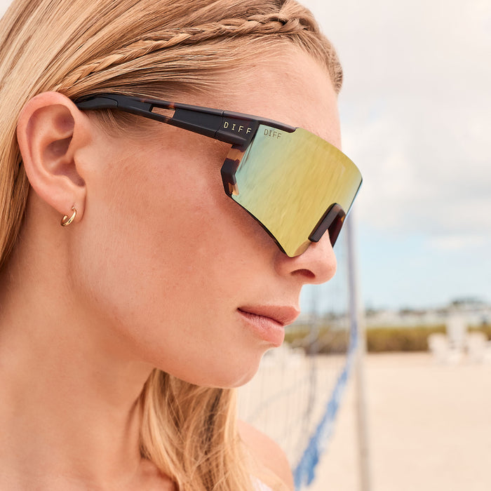 lifestyle - blonde female about to play beach volley ball while wearing diff sport heat shield sunglasses with a sport tortoise frame and orange flash polarized lenses close up view