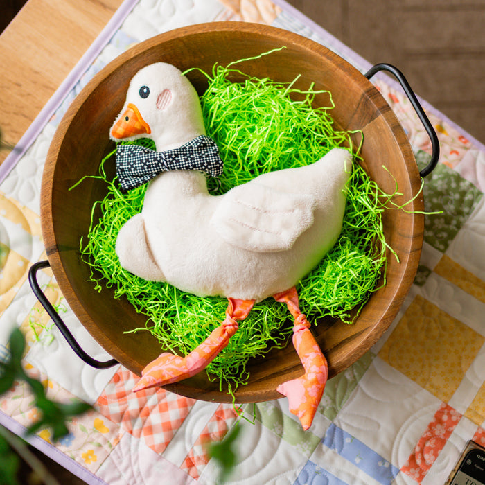 Decorative goose figurine with a bow tie in a wooden bowl on a patterned tablecloth.