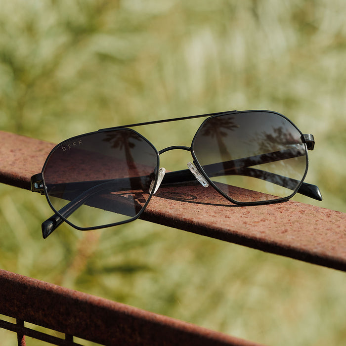 still life-  jojo fletcher x diff eyewear joelle navigator sunglasses with a black metal frame and grey gradient lenses set on a railing
