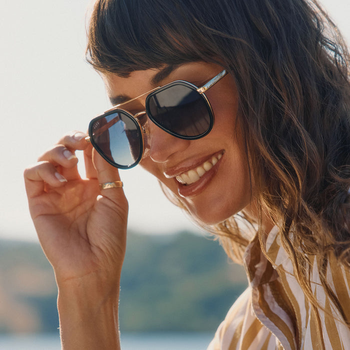 lifestyle - female at the lake wearing diff eyewear featuring the joey round sunglasses with a black & gold metal frame with grey gradient polarized lenses angled view
