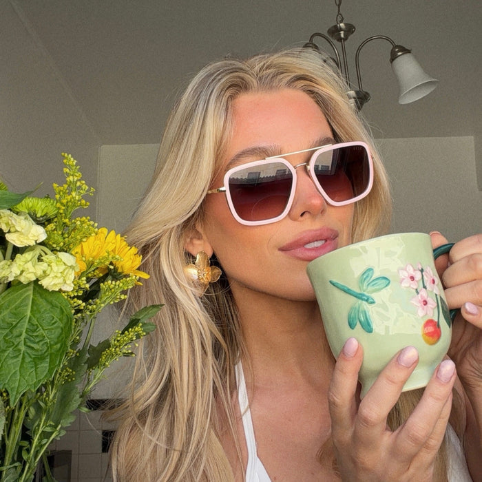 lifestyle - shelby hunstad blonde influencer wearing baby pink navigator sunglasses with pastel painted tea mug and spring flowers