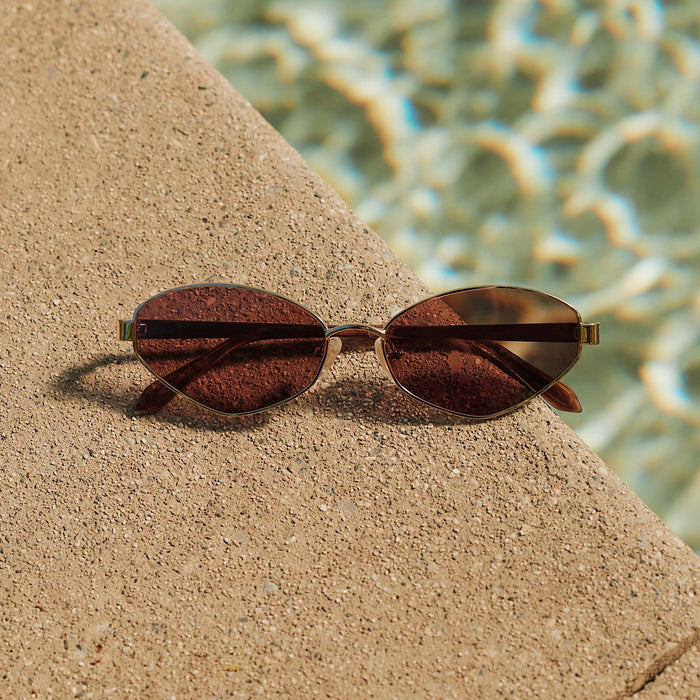 still life -  jojo fletcher x diff eyewear jordy cat eye sunglasses with a gold metal frame with horn tortoise temples and brown lenses laying next to a pool