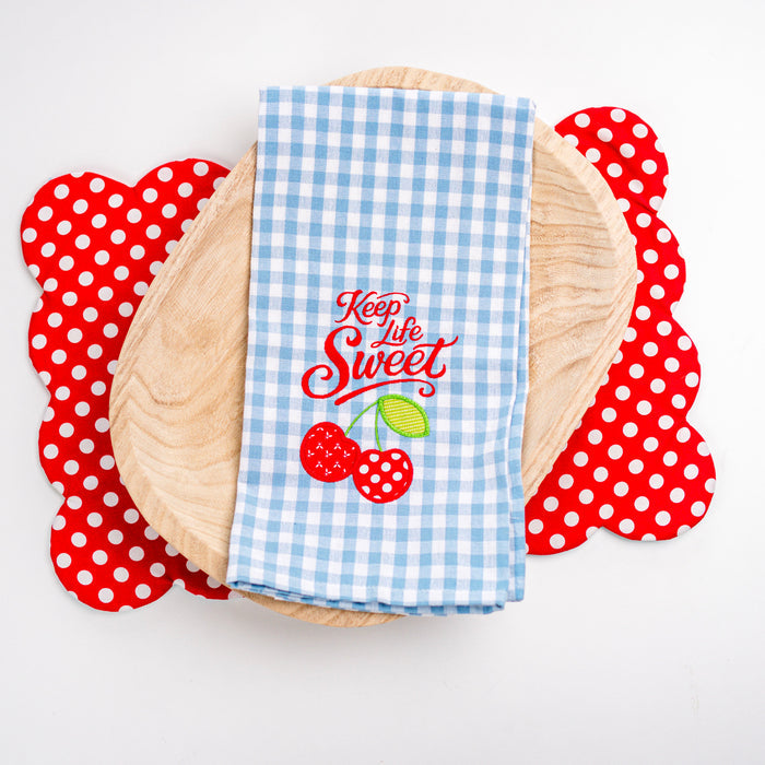 Blue checkered towel with 'Keep Life Sweet' text and cherry design on a wooden plate with red polka dot coasters.