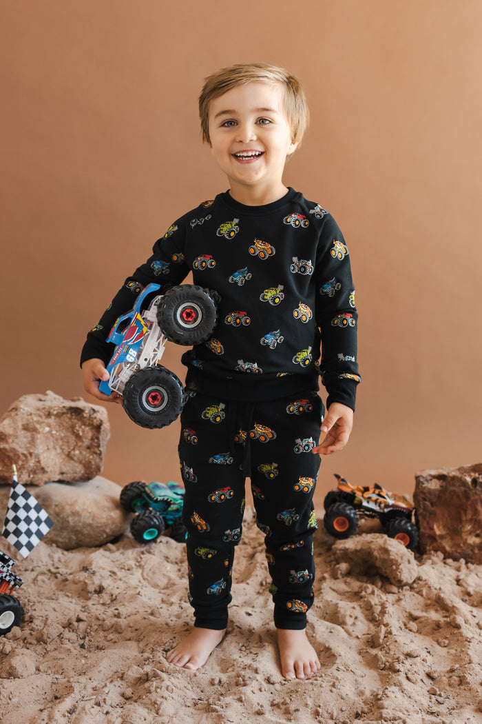 Toddler wearing Black Long Sleeve Jogger Set in Monster Truck print holding a truck standing on sand and rock setup