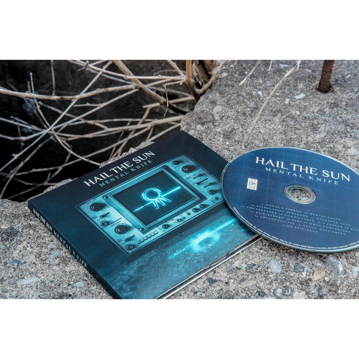 CD cover is a picture of a screen with buttons on either side. The screen has the Hail the Sun logo printed on it in glowing blue color. The logo from the screen is being reflected onto the floor. Above the screen has "HAIL THE SUN METAL KNIFE" printed. CD and cover is placed on the ground outside with sticks in the background.