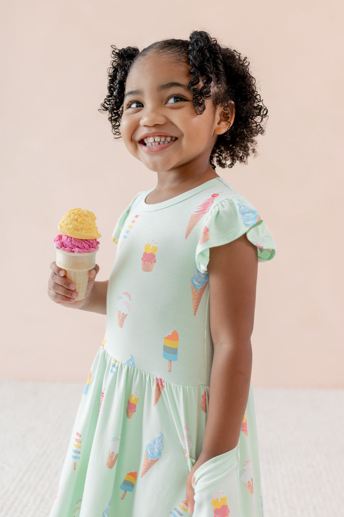 Side view of child in Pocket Dress with Mint Ice Cream print holding an ice cream on a beige background