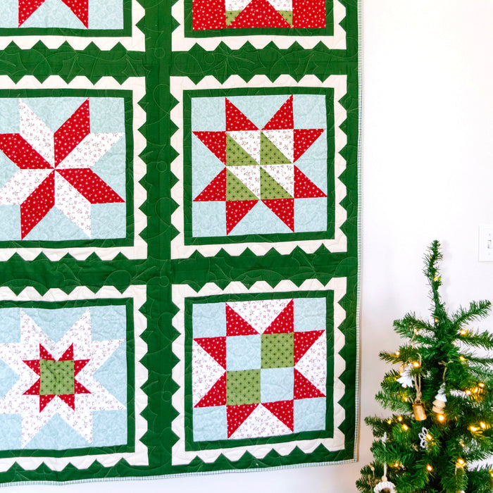 A handmade quilted wall hanging with six Christmas star and snowflake patterns in red, green, and light blue, displayed on a black metal rod against a white wall next to a decorated Christmas tree.