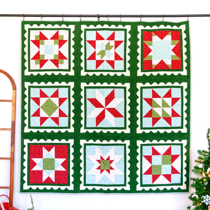 A large quilted wall hanging with a 3x3 grid of holiday starburst and snowflake-style blocks, featuring a dark green border and red, white, and light blue patterned squares, displayed on a black rod against a white wall with a wooden ladder and a small Christmas tree nearby.