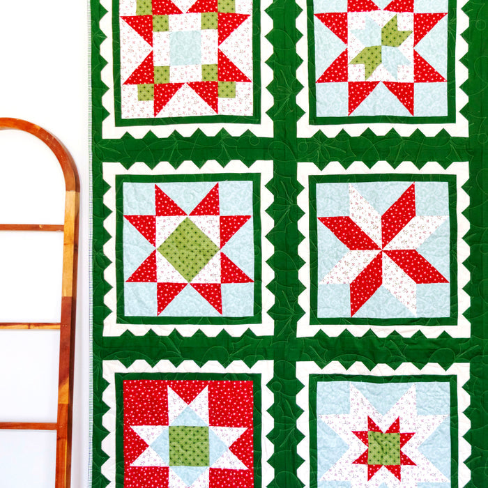 A handmade quilt with a 3x3 grid of holiday starburst patterns, featuring red, green, and white fabrics with a dark green border, hanging on a white wall next to a wooden ladder and a small Christmas tree.