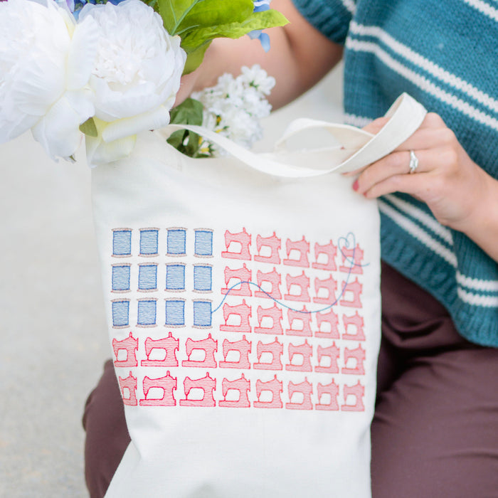 Person holding a tote bag with a pattern, surrounded by flowers and wearing a blue sweater.