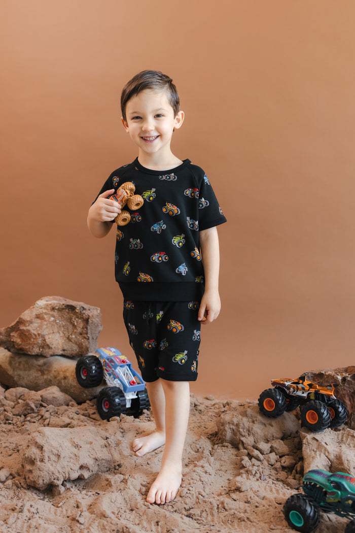 Boy smiling in Black Short Sleeve Jogger Set in Monster Truck print holding toy truck while standing on sand and rocks with toy trucks